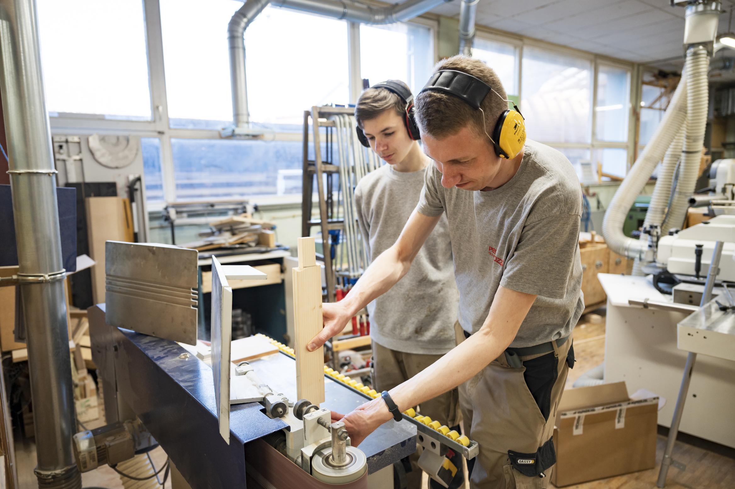 zwei junge Männer in Arbeitskluft und mit Gehörschutz arbeiten an einer Schleifmaschine in einer Schreinerei