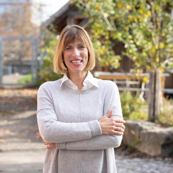 Frau mit kurzem blondem Haar, grauem Pullover und weißer Bluse steht lächelnd mit verschränkten Armen in einem herbstlichen Innenhof.