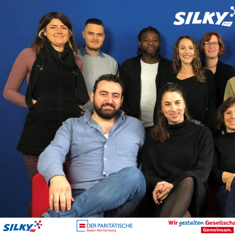 Eine Gruppe junger Menschen vor einer blauen Wand. 8 Personen stehend, 4 Personen sitzend davor auf einer roten Couch