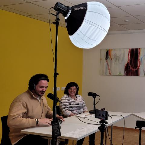 vier Teilnehmer*innen sitzen an Tischen und produzieren einen Podcast. Der Raum ist mit Scheinwerfern erleuchtet.