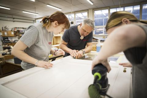 3 Personen beugen sich über eine Holzarbeitsplatte, eine Person sägt mit einer Stichsäge in einer Werkstatt