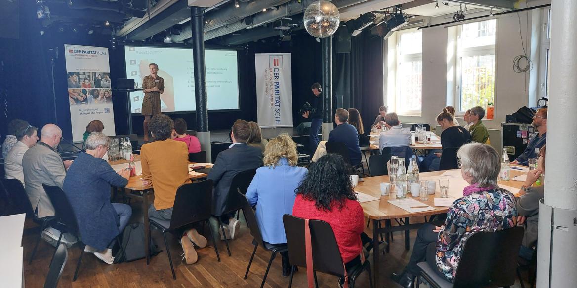 Frau auf der Bühne hält einen Vortrag und die Zuschauer an den Tischen hören zu