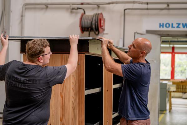 zwei Männer arbeiten an einem Holzschrank in einer Werkstatt