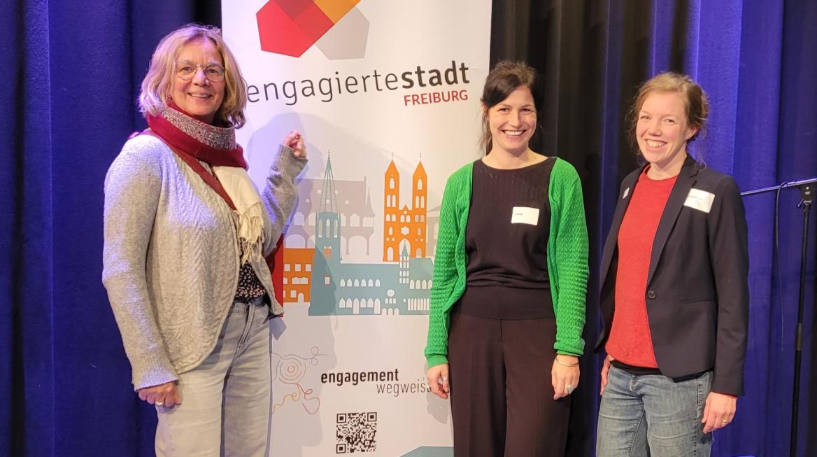 Drei Frauen stehen an einem Roll-up auf dem "engagierte Stadt Freiburg" steht