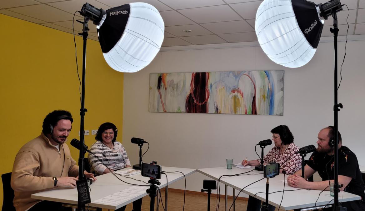vier Teilnehmende sitzen bei einem Podcast. Der Raum wird von Scheinwerfern erleuchtet  und es sind Mikrofone auf dem Tisch aufgebaut.