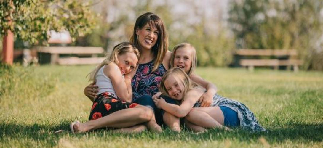 eine Frau sitzt mit 3 Mädchen im Arm lachend auf einer Sommerwiese