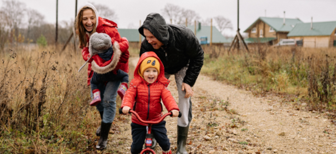 Mutter, Vater, Kinder auf einem Feldweg im Regen, ein Kind auf einem Fahrrad, es wird vom Vater angeschoben-