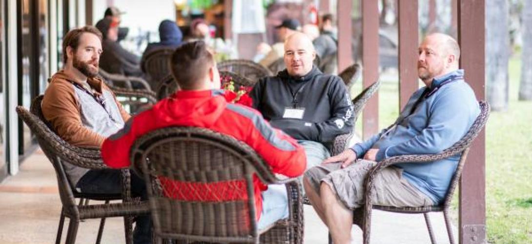 kleine Gruppe von m&auml;nnlich gelesenen Personen sitzt im Kreis um einen Tisch und unterh&auml;lt sich