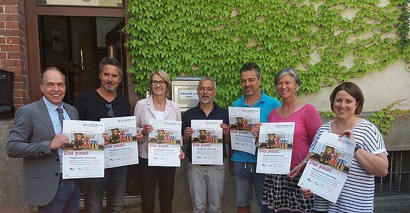 ier Männer und dreu Frauen stehen nebeneinander im Halbkreis vor einer mit Efeu bewachsenen Hauswand und halten das Kampagnenplakt hoch mit der Aufschrift "Das passt. Kindern Familie ermöglichen".