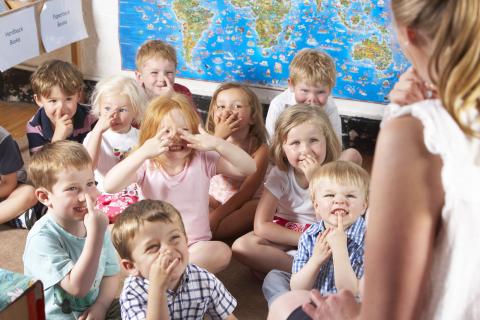 Zehn Kinder /sechs Jungen und vier Mädchen) sitzen im Schneidersitz auf dem Boden im Klassenzimmer und zeigen mit dem Finger auf ihre Nase. Im Vordergrund steht die Lehrerin und macht die Bewegung vor. Im Hintergrund hängt an der Wand eine Weltkarte.