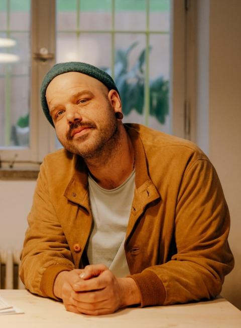 Bild von jungem Mann mit hellbraunem Hemd, grauer Mütze und Oberlippenbart. 