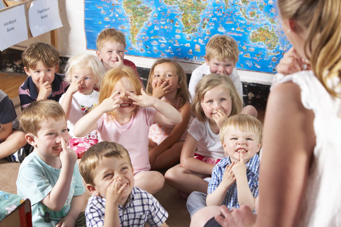 Kleine Kinder deuten gemeinsam mit junger Frau mit Hand auf Mund, Nase oder Stirn
