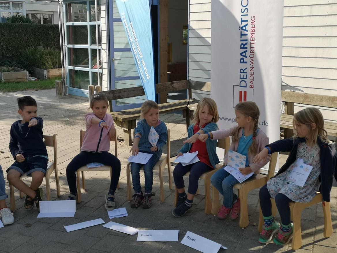 Kleine M&auml;dchen und Jungs sitzen im Stuhlkreis und strechen ihren Arm mit dem Dankesch&ouml;narmb&auml;ndchen raus