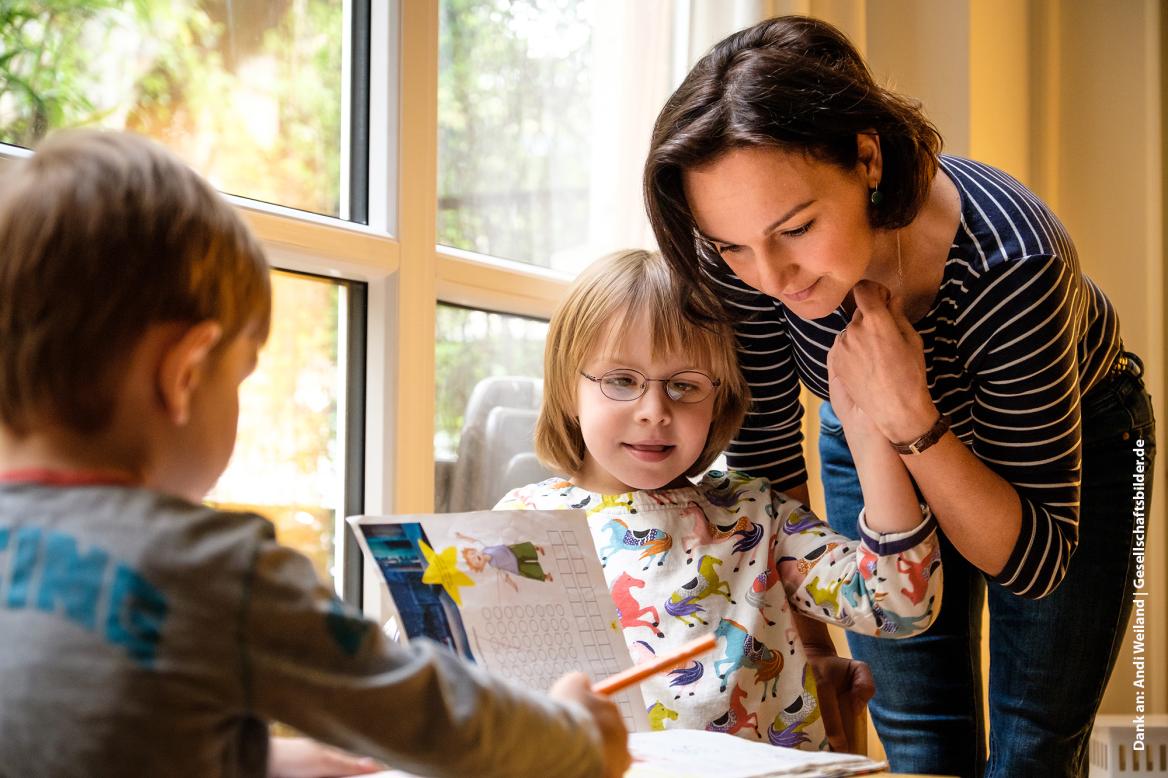 Zwei Kinder am Tisch machen Hausaufgaben. Mutter sieht sich die Aufgaben an.