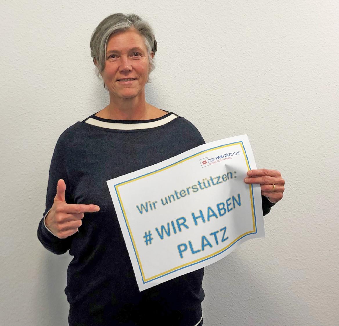 Stehende Frau h&auml;lt ein Plakat in einer Hand und deutet mit der anderen auf die Aufschrift #Wir haben Platz